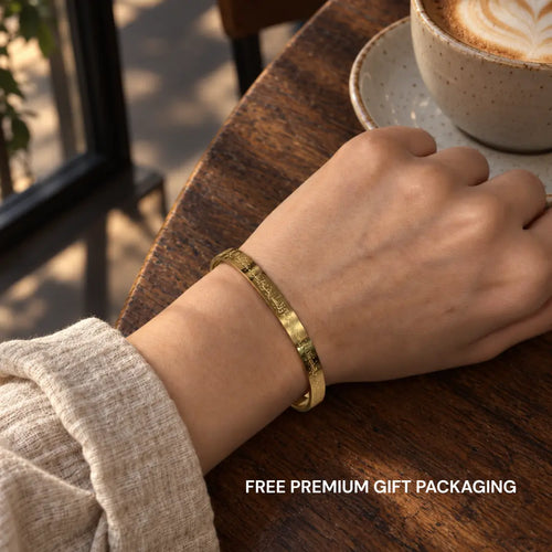 A gold engraved bangle bracelet worn on a person's wrist, resting on a wooden table next to a cup of latte. The text 'FREE PREMIUM GIFT PACKAGING' is visible at the bottom right.