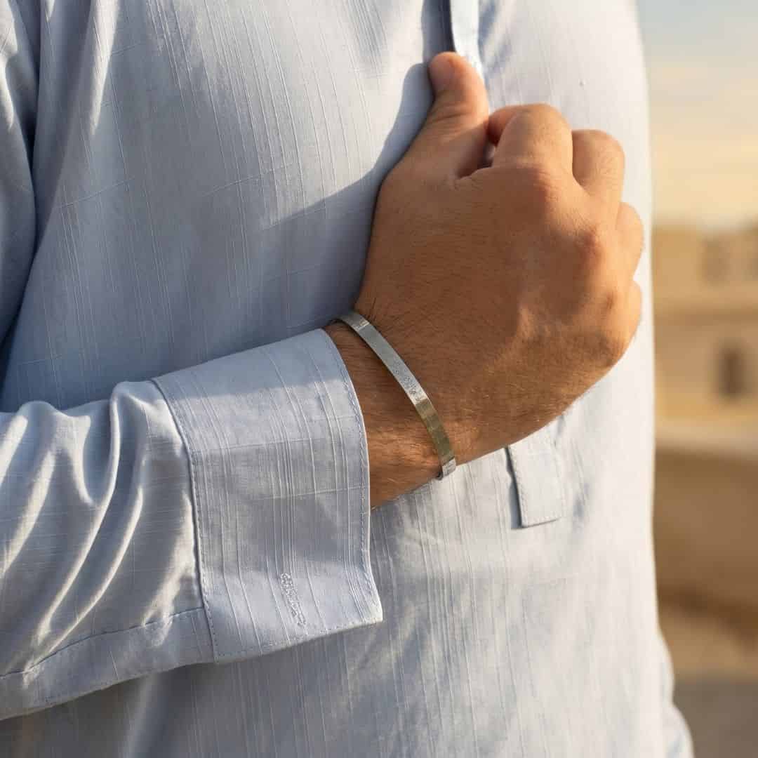 A person wearing a slim silver bangle bracelet, shown from the side with their hand resting on a light-colored fabric surface in a softly lit indoor setting.