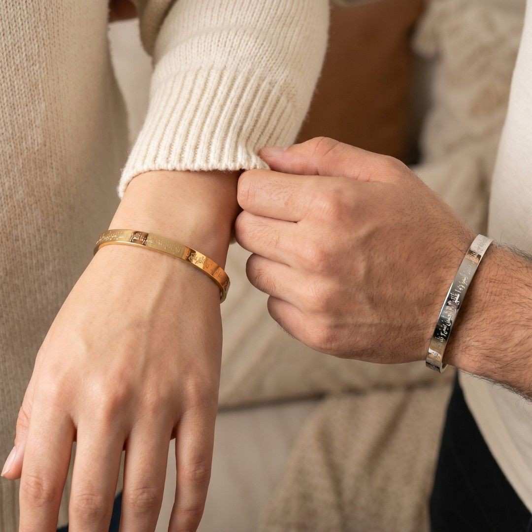 A close-up lifestyle shot of a person adjusting a gold bangle bracelet on their wrist while wearing a matching silver bangle on the other wrist. The background features a soft beige knit sweater.