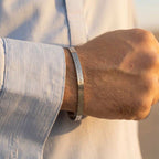 A close-up of a man's wrist wearing a polished silver metal cuff bracelet engraved with the word 'LOVE' in vertical lettering, wearing a light blue linen shirt.