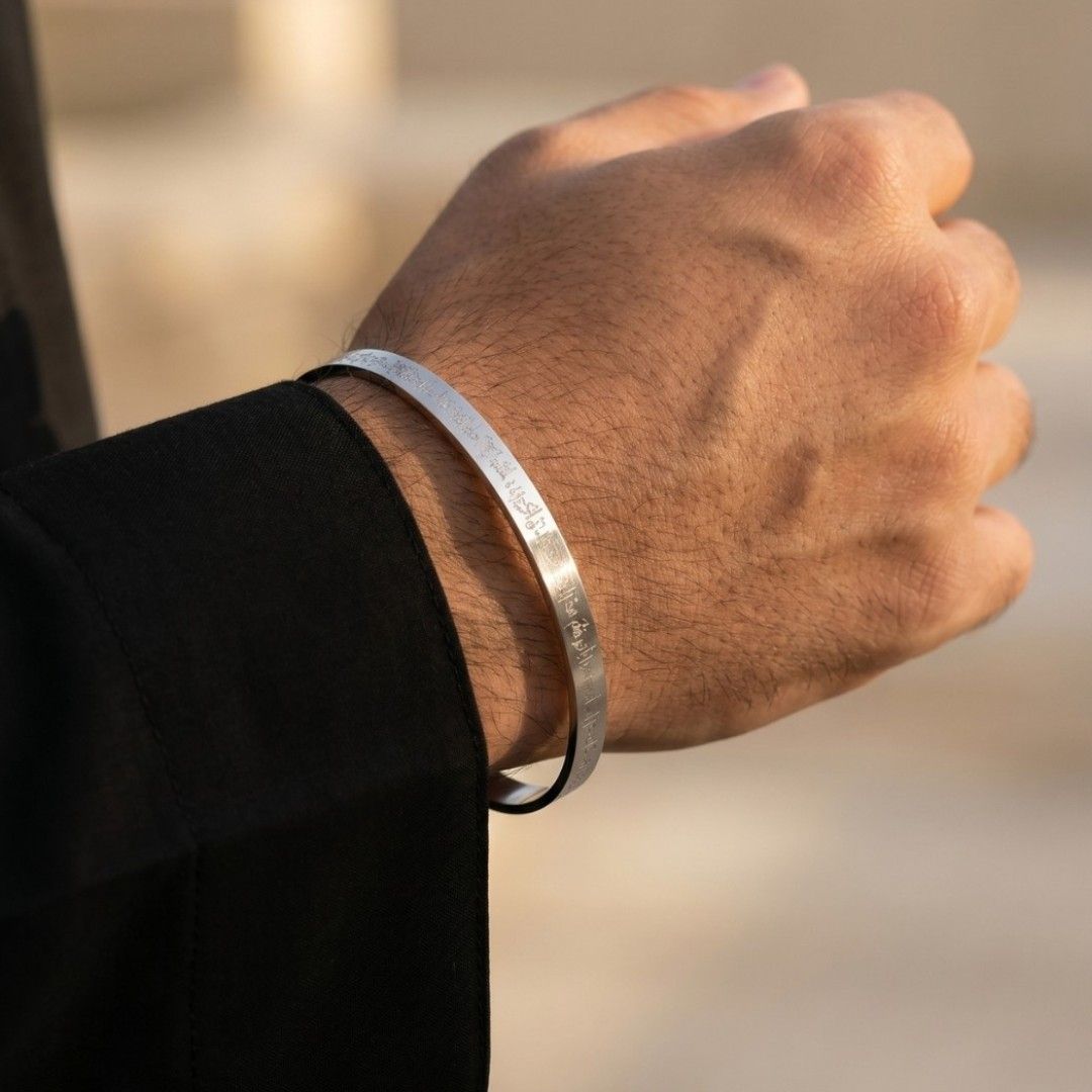 A close-up of a person's hand wearing two slim silver metal bracelets, one engraved with 'JESUS IS MY SAVIOR' and the other with 'JESUS IS MY GOD', set against a warm, blurred background.