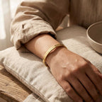 A close-up of a person's hand and forearm resting on a textured beige cushion, wearing a thin gold bangle bracelet with engraved lettering.