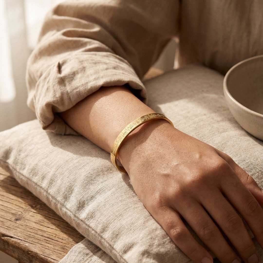 A close-up of a person's hand and forearm resting on a textured beige cushion, wearing a thin gold bangle bracelet with engraved lettering.