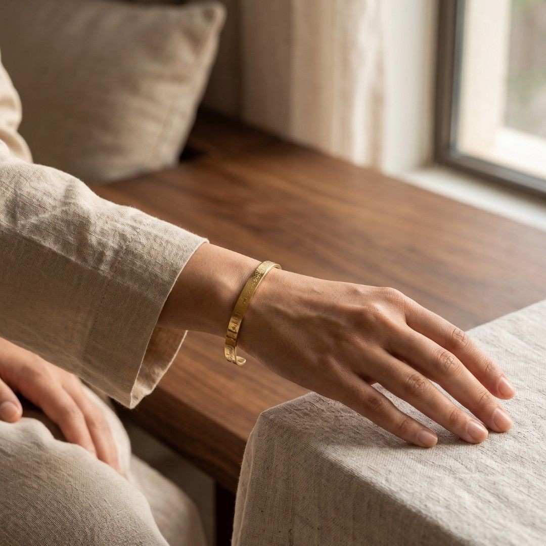 A person wearing a slim gold bangle bracelet, shown from the side with their hand resting on a light-colored fabric surface in a softly lit indoor setting.