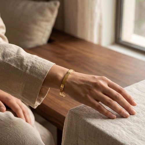 A person wearing a slim gold bangle bracelet, shown from the side with their hand resting on a light-colored fabric surface in a softly lit indoor setting.
